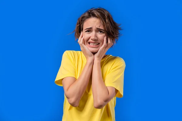 Woman afraid of something and looks into camera with big eyes full of pain on blue background. Phobia, trouble, panic concept. High quality photo