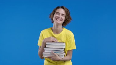 Zeki bir öğrenci kadın üniversite kütüphanesinden mavi arka planda bir yığın kitap tutuyor. Mutlu kız gülümser, mezun olduğu için mutludur. Yüksek kalite 4k görüntü