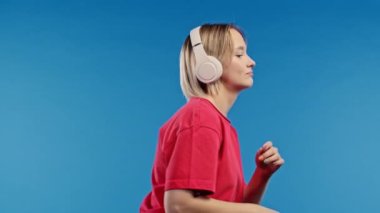 Müzik dinleyen pozitif sarışın kadın, mavi stüdyo arka planında kulaklıklarla dans etmekten hoşlanıyor. Radyo, kablosuz modern ses teknolojisi, çevrimiçi çalar. Yüksek kalite 4k görüntü