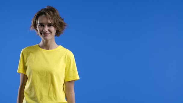 青い背景に黄色の服を着た美しい女性 コピースペース カメラを探している若い女性 短い流行の髪型 陽気な笑顔 高品質4K映像 — ストック動画
