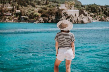 Çizgili gezi kıyafeti giymiş, hasır şapkalı bir kadın. Taş plajda duran bir kadın. Teal denizi arka planı. Güney Avrupa Riviera, seyahat yeri, lüks tatil konsepti. Yüksek kalite fotoğraf