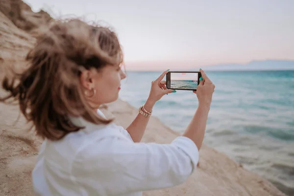Young woman filming video with mobile phone on beach near blue sea. Girl using smartphone, taking photos for social app, enjoying vacation time. Memory concept. High quality photo
