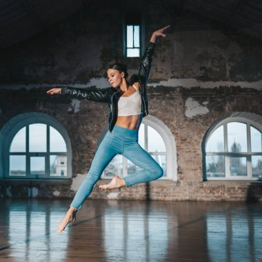 Young woman jumping in casual style - jeans and leather jacket doing ballet in old studio. Attractive ballerina practices in choreography alone. High quality photo clipart