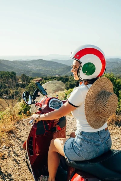 Woman sitting on retro scooter or motor bike in red color, traveling in mountains on island. Tourist in helmet enjoying nature of amazing destination. Summer landscape. High quality photo