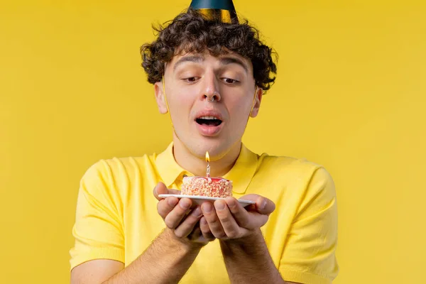 Happy birthday man making wish - blowing candle on cake. Curly haired guy laughs, celebrating anniversary. Young teenager on yellow background. High quality photo