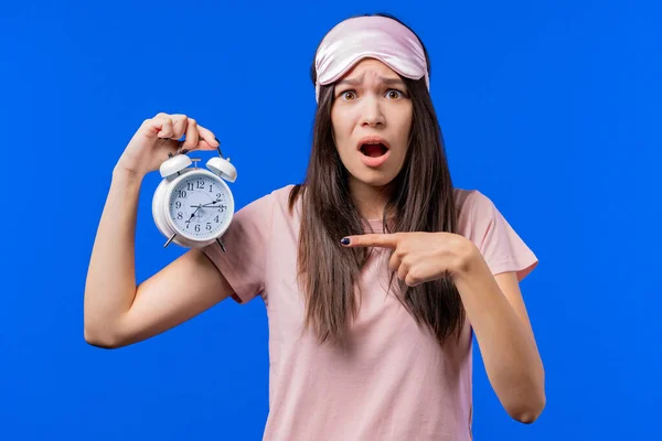 Woken up by alarm clock sleepy woman holding it in hand. Blue background. Early 7 o\'clock in morning. Lazy teen girl didn\'t get enough sleep, concept of passing time. High quality photo