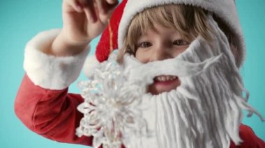 Noel Baba kostümlü sevimli bir çocuk, yapay sakallı, kar tanesi Noel ağacı oyuncağıyla oynayan. Şenlik havası. Mutlu çocukluk, çocuklar, komik çocuk. Yüksek kalite 4k görüntü