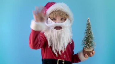 Noel Baba kostümlü arkadaş canlısı küçük Noel çocuğu el sallıyor. Merhaba. Selamlar, mavi arka plandaki kameraya merhaba deyin. Yapay sakallı mutlu duygusal çocuk. Yüksek kalite 4k görüntü