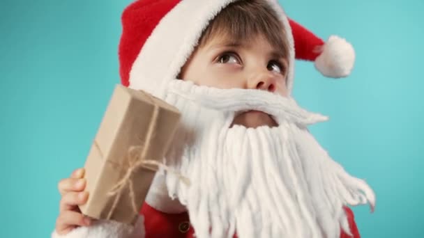 弓でギフトボックスを保持髭とサンタの衣装で興奮した少年 子供の中に何があるのだろうか プレゼント付きの幸せな子供 青いクリスマスの背景 高品質4K映像 — ストック動画