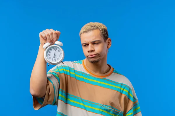 Woken up by alarm clock sleepy man holding it in hand. Blue background. Early 6 o\'clock in morning. Lazy guy didn\'t get enough sleep, concept of passing time. High quality photo