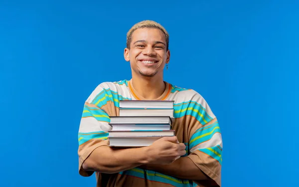Young man student holds stack of university books from college library on blue background. Happy guy smiles, he is happy to graduate, education abroad concept. High quality photo