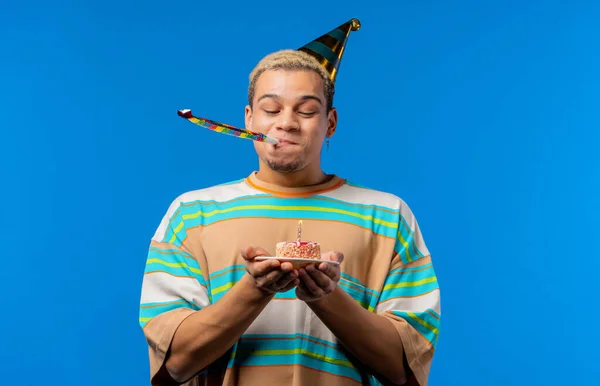 Happy birthday man making wish - blowing candle on cake. Curly haired guy laughs, celebrating anniversary. Young teenager on blue background. High quality photo
