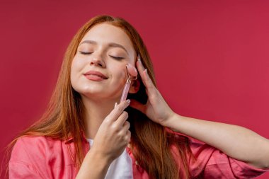 Young woman doing face massage with rose quartz stone roller on viva magenta background. Facial self care, beauty rituals, cosmetology, anti aging and anti-wrinkle treatment. High quality photo