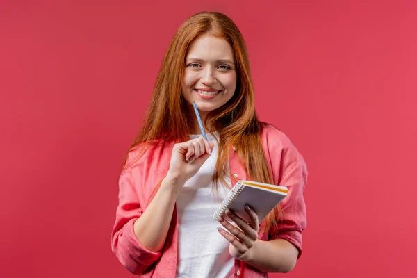 Young woman writing notes in planner with pen. Ginger girl thinking about future plans and to-do list in notebook for week, month, year. Keeping personal diary on pink background. High quality photo
