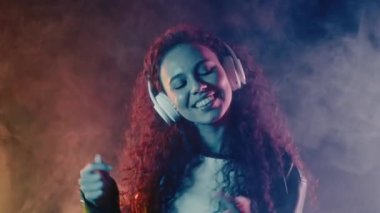 Curly haired woman listening music, singing, enjoying dance with headphones in smoke. Radio, wireless modern sound technology, online player. High quality 4k footage