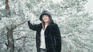 Stylish woman in fluffy bucket hat pulls on branch of tree, snow flies on her and she is happy rejoices, winter and cold weather. Slow motion. High quality 4k footage