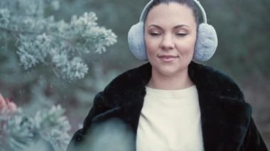 Stylish woman in fluffy ear muffs walking in winter frozen forest. Lady touches branch of pine, enjoys nature. Snowflakes fly by. High quality 4k footage