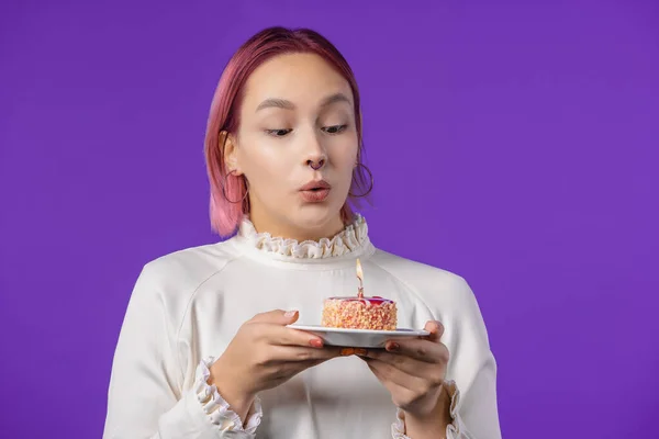 Birthday woman making wish - candle on cake. Girl smiling, celebrating anniversary. Young stylish lady with pink dyed hairstyle on violet background. High quality photo
