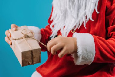 Hands of boy in Santas costume pulling on string of wrapped gift box. Child wonders what is inside. Happy kid with present. Blue Christmas background. High quality photo
