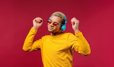 Positive american man listening music, enjoying dance with headphones on red studio background. Radio, wireless modern sound technology, online player. High quality photo