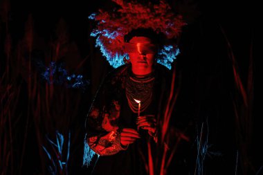 Geceleri kamışında mum olan Ukraynalı bir kadın. Gözlerinin etrafındaki kırmızı kurdele, oyuncak bebeklerdeki gibi. Geleneksel kostüm. Sembolizm, antik büyü, doğanın gücü, kostüm oyunu 