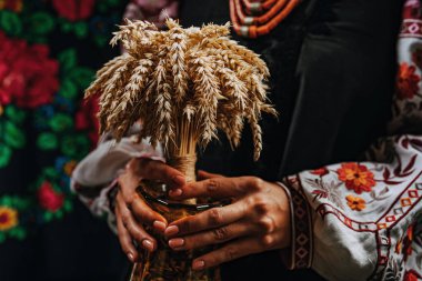 Ukrainian woman in traditional costume with bouquet Didukh on colorful bukovyna ornaments carpet. Christmas decoration, made from sheaf of wheat.  clipart