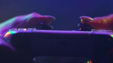 Kadın oyuncu konsolda online video oyunu oynuyor. Neon renkli ışığın altında kablosuz kontrolör. Kız, joypad kullanarak düğmelere basıyor. Hobi, e-spor, makro görüş