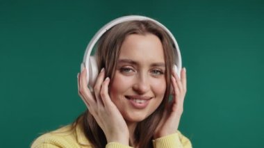 Müzik dinleyen mutlu bir kadın, yeşil stüdyo arka planında kulaklıklarla dans ediyor. Radyo, kablosuz modern ses teknolojisi, çevrimiçi oynatıcı.