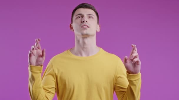 紫色の背景に交差指で祈る若い男 男は誰かが欲望を満たし 試験で運を祈ることを懇願します 高品質4K映像 — ストック動画