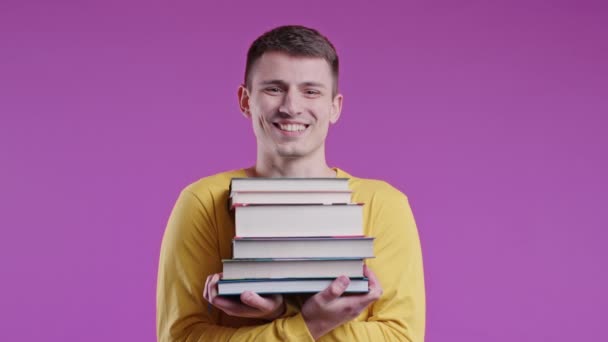 若い男性学生はピンクを背景に大学図書館から大学図書を積み上げています 幸せな男は笑顔 彼は海外で卒業し 教育に満足している — ストック動画