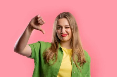 Portrait of unhappy european woman condemns with sign of dislike. Young millennial lady expressing discontent with showing thumbs-down gesture on pink studio background. clipart
