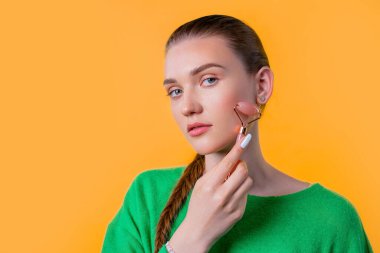 Sarı arka planda gül kuvars taşıyla yüz masajı yapan genç bir kadın. Yüz bakımı, güzellik ritüelleri, kozmetoloji, yaşlanma karşıtı ve kırışıklık önleyici tedavi