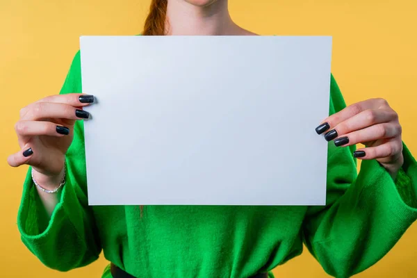 stock image Young woman holding horizontal white a4 paper poster. Copy space. Smiling girl in green sweater on yellow background.