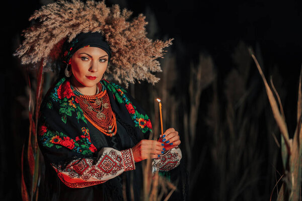 Mysterious ukrainian woman with candle, she in traditional costume outdoors at night. Lady in authentic jewelry. Beauty of Ukraine, ethnic clothing, details, light, faith hope
