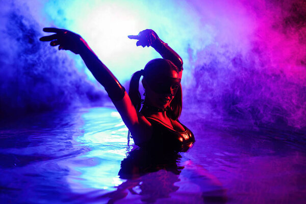 Sensual woman in leather BDSM mask dancing in swimming pool, neon light. Attractive chick enjoying party time. Adult entertainment, masquerade. Dangerous lady. High quality photo