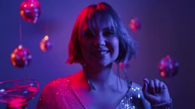 Şampanya bardağıyla dans eden güzel bir kadın, yeni yıl partisi, neon renkli bir ışık. Kutlama, gece kulübü, boyalı hippi pembe saç, parlak elbise. Yüksek kalite 4k görüntü