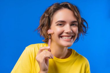 Mavi arka planda gül kuvars taşıyla yüz masajı yapan genç bir kadın. Yüz bakımı, güzellik ritüelleri, kozmetoloji, yaşlanmayı önleme ve kırışıklık önleme tedavisi. Yüksek kalite fotoğraf