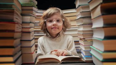 Gözlüklü şirin sarışın okul çocuğu kütüphanede kitap yığınları arasında ilginç kitaplar okuyor. Eğitim konsepti, çocuk zevki zamanı. Yüksek kalite 4k görüntü