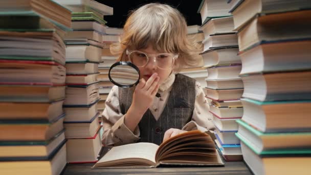 真剣な小さな研究者の少年は 図書館でグラスを拡大した本を読む 楽器で知識を学ぶメガネでかわいいプリスクーラー 高品質の4K映像 — ストック動画