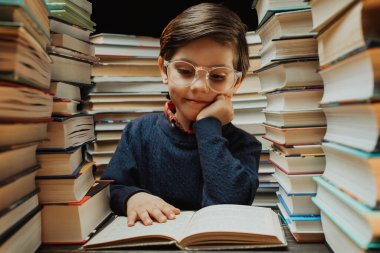Gözlüklü şirin öğrenci kütüphanede kitap yığınları arasında ilginç kitaplar okuyor. Eğitim konsepti, hazırlık ya da ilkokul. Yüksek kalite