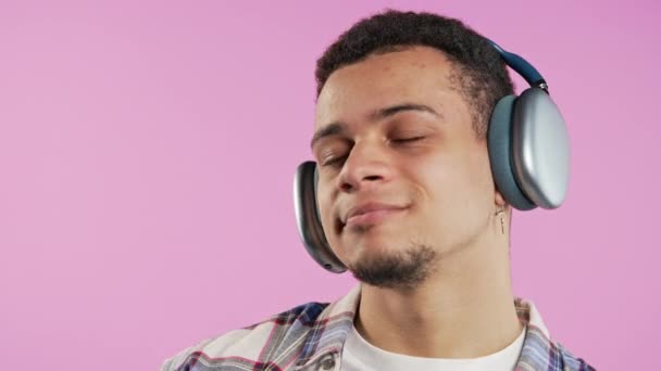 ポジティブなミックスレースマンは音楽を聴き ピンクのスタジオの背景にトレンディなモダンなヘッドフォンでダンスを楽しんでいます ラジオ ワイヤレス現代サウンド技術 オンラインプレーヤー 高品質4Kについて — ストック動画