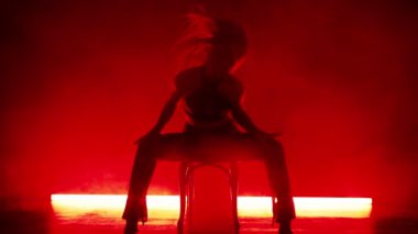 Ateşli kadın seksi saç hareketleri yapıyor, kırmızı neon ışıklı sahnede sandalyeye oturmuş başını sallıyor. Baştan çıkarıcı özel dans. Duman içindeki bayan dansçı. Yüksek kalite 4k görüntü