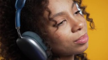Müzik dinleyen Afro-Amerikan kadın, sarı stüdyo arka planında kulaklıklarla eğleniyor. Radyo, kablosuz modern ses teknolojisi, çevrimiçi çalar. Yüksek kalite 4k