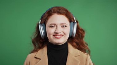 Uzun kızıl saçlı, müzik dinleyen, yeşil stüdyo arka planında kulaklıkları olan pozitif öğrenci bir kadın. Radyo, kablosuz modern ses teknolojisi, çevrimiçi çalar. Yüksek kalite 4k