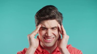 Baş ağrısı çeken genç bir adam, stüdyo portresi. Adam ellerini başının üstüne koyuyor, mavi arka planda izole edilmiş. Sorunlar, ilaç, hastalık kavramı