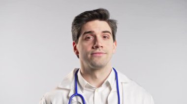 Yakışıklı doktor gözünü kırpıyor ve kameraya gülümsüyor. Beyaz hastane stüdyosunda arka planda biri var. Yüksek kalite 4k görüntü