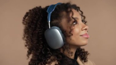 Mutlu Afro-Amerikan kadınları kafayı buluyor, müzik dinliyor, bej renkli stüdyo arka planında kulaklıklarla dans ediyor. Radyo, kablosuz modern ses teknolojisi, çevrimiçi çalar. Yüksek kalite 4k