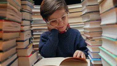 Gözlüklü şirin öğrenci kütüphanede kitap yığınları arasında ilginç kitaplar okuyor. Eğitim konsepti, hazırlık ya da ilkokul. Yüksek kalite 4k görüntü