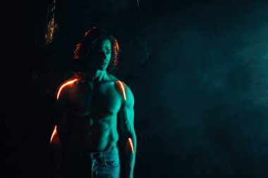 Uzun saçlı, çıplak vücutlu yakışıklı bir adam. Renkli ışıklandırma, lazer ışığı, neon duman klubü altında çıplak karın kası portresi. Projeksiyon illüzyon haritası. Fütürist model.