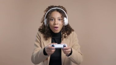 Oldukça zoomer genç Afro-Amerikan kadın TV 'de heyecan verici video oyunu oynuyor. Oyun kolu bej renkli stüdyo arka planında izole edilmiş. Modern teknolojiyi kullanarak. E-spor, hobi, akış